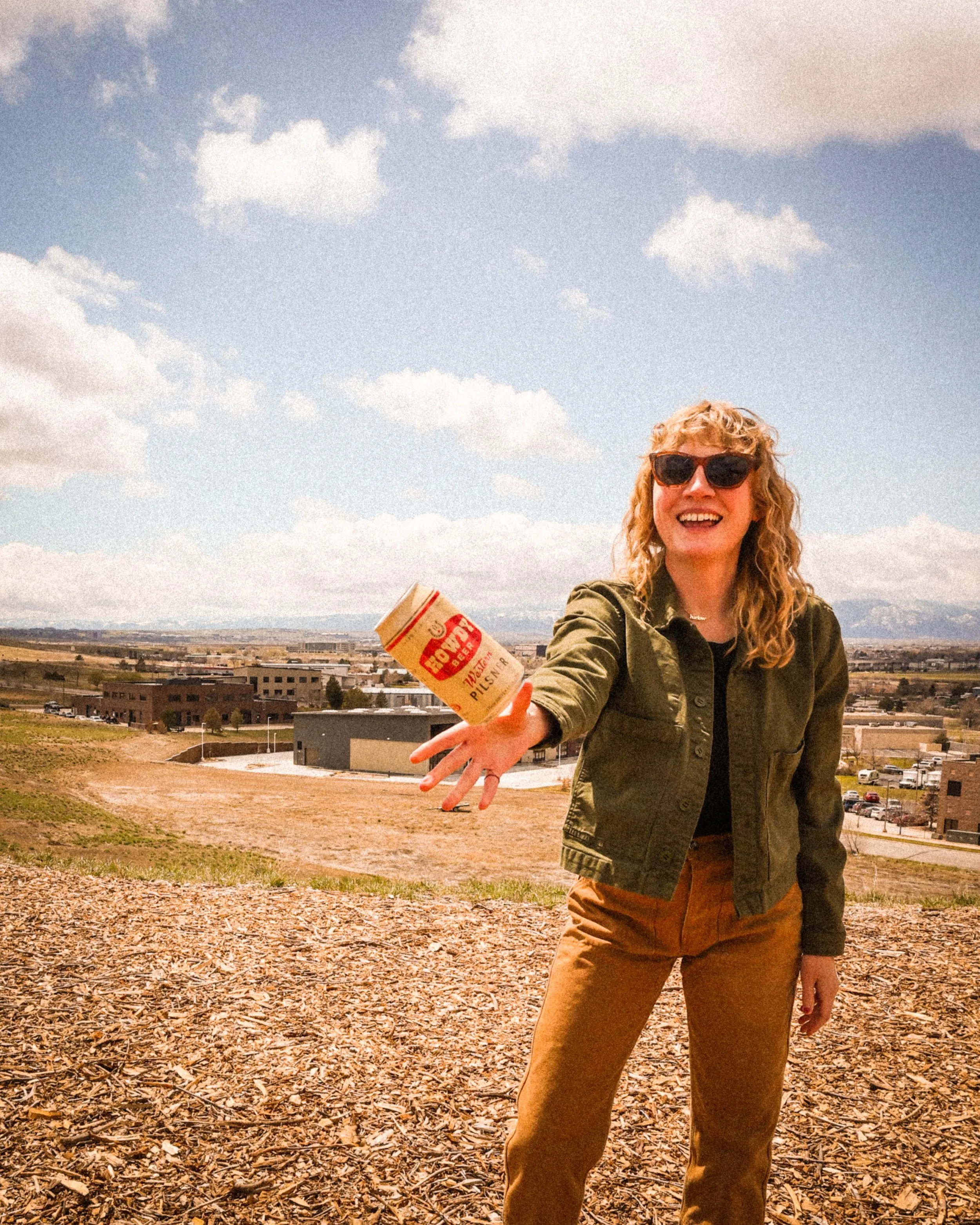 Smiling person tossing a Howdy Western Pilsner can toward the camera with mountains behind