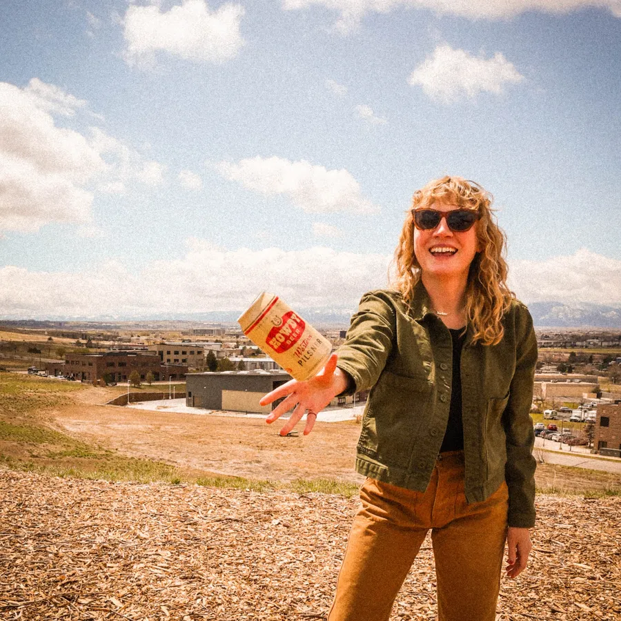 Smiling person tossing a Howdy Western Pilsner can toward the camera with mountains behind