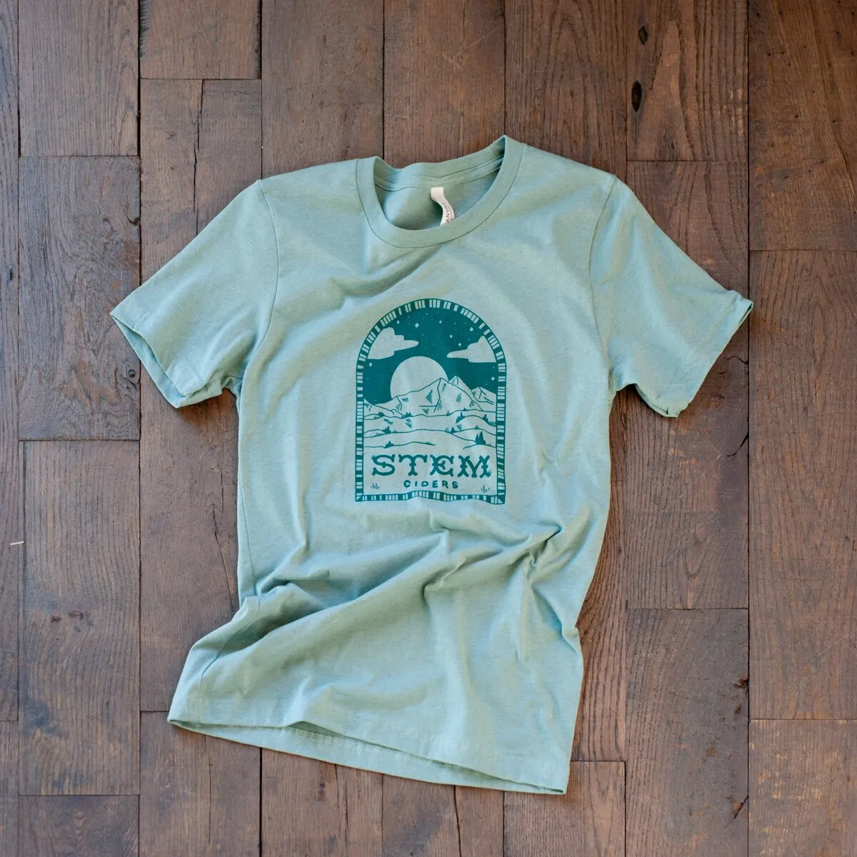Sage green Stem Ciders t-shirt laid on a wood floor