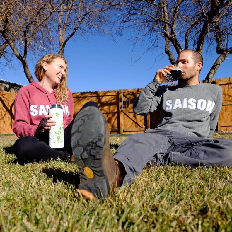 Two people in pink and gray Saison sweatshirts sitting in grass